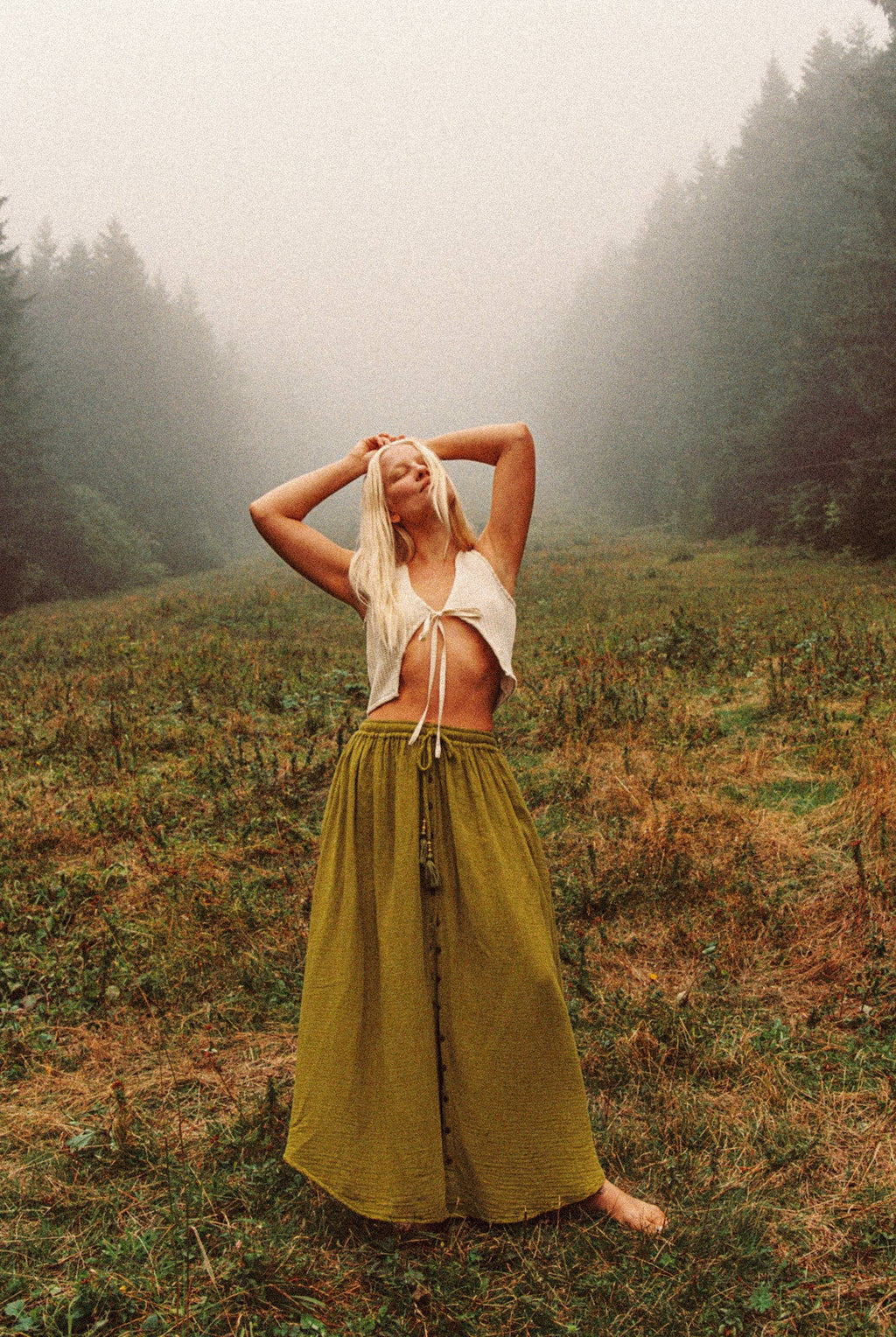 Woman in a white top and long green skirt standing in a foggy forest clearing.