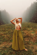 Woman in a white top and long green skirt standing in a foggy forest clearing.