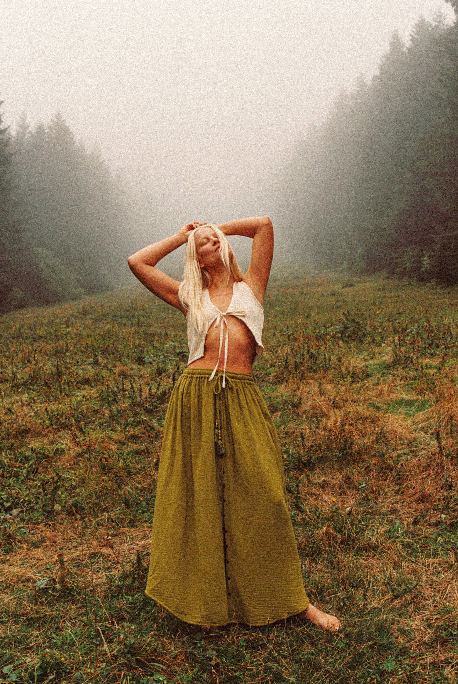 Woman in a white top and long green skirt standing in a foggy forest clearing.