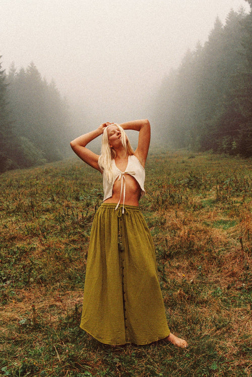 Woman in a white top and long green skirt standing in a foggy forest clearing.