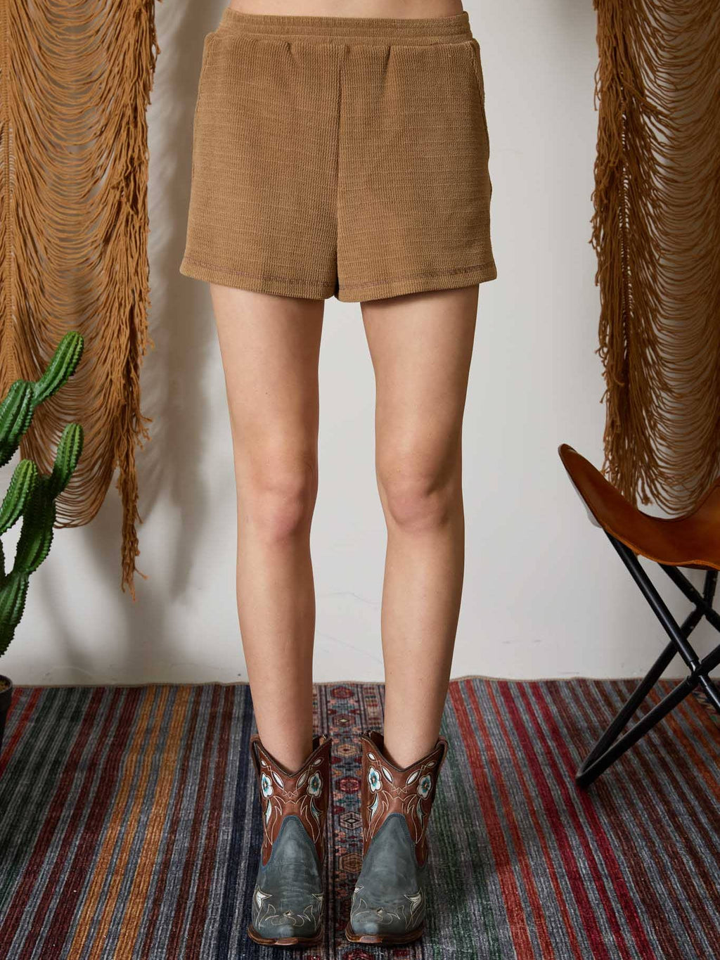 A person shown from the waist down wearing tan corduroy shorts and brown western boots, standing on a patterned rug with fringe wall hangings and a chair in the background.
