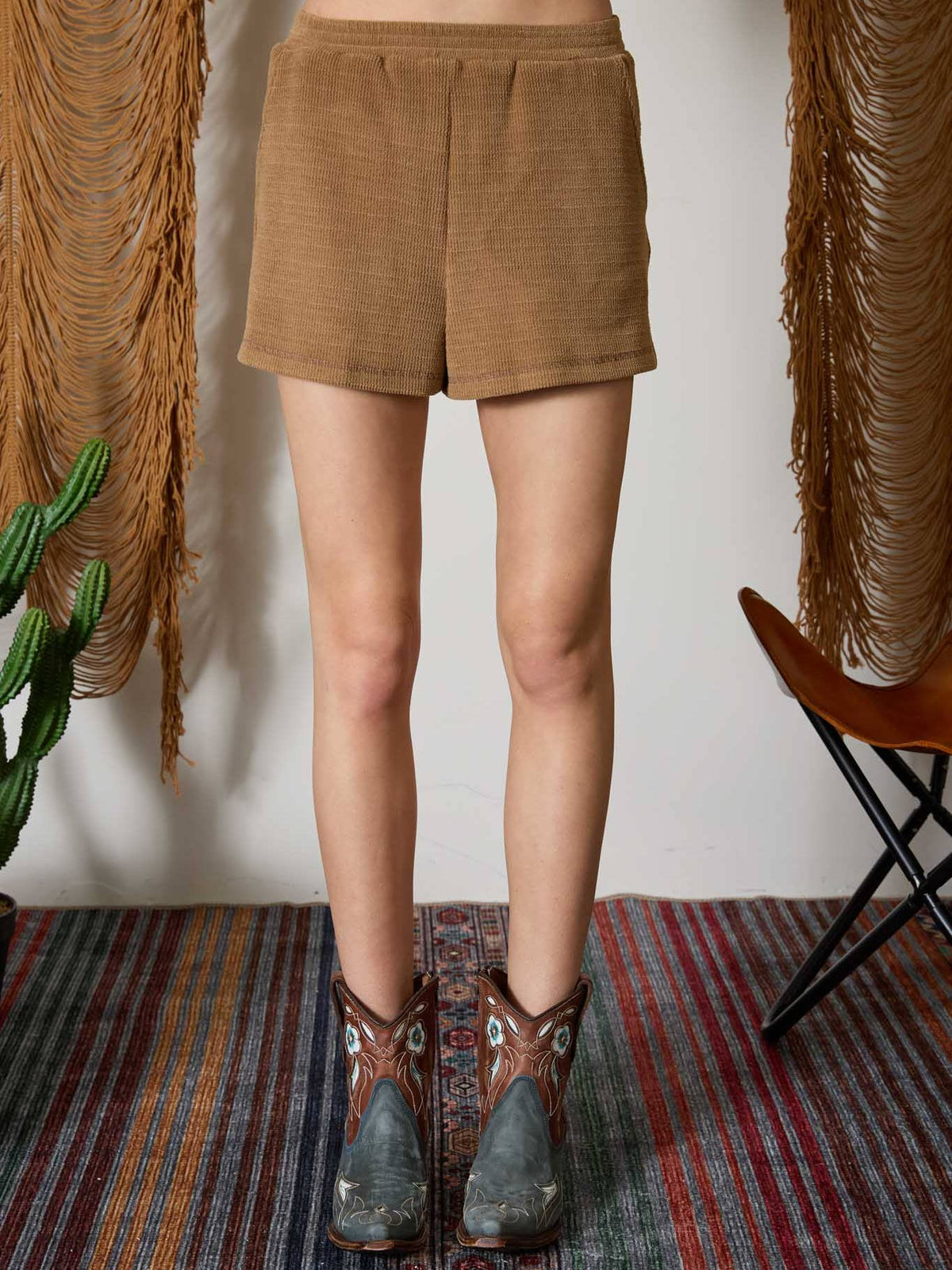 A person shown from the waist down wearing tan corduroy shorts and brown western boots, standing on a patterned rug with fringe wall hangings and a chair in the background.