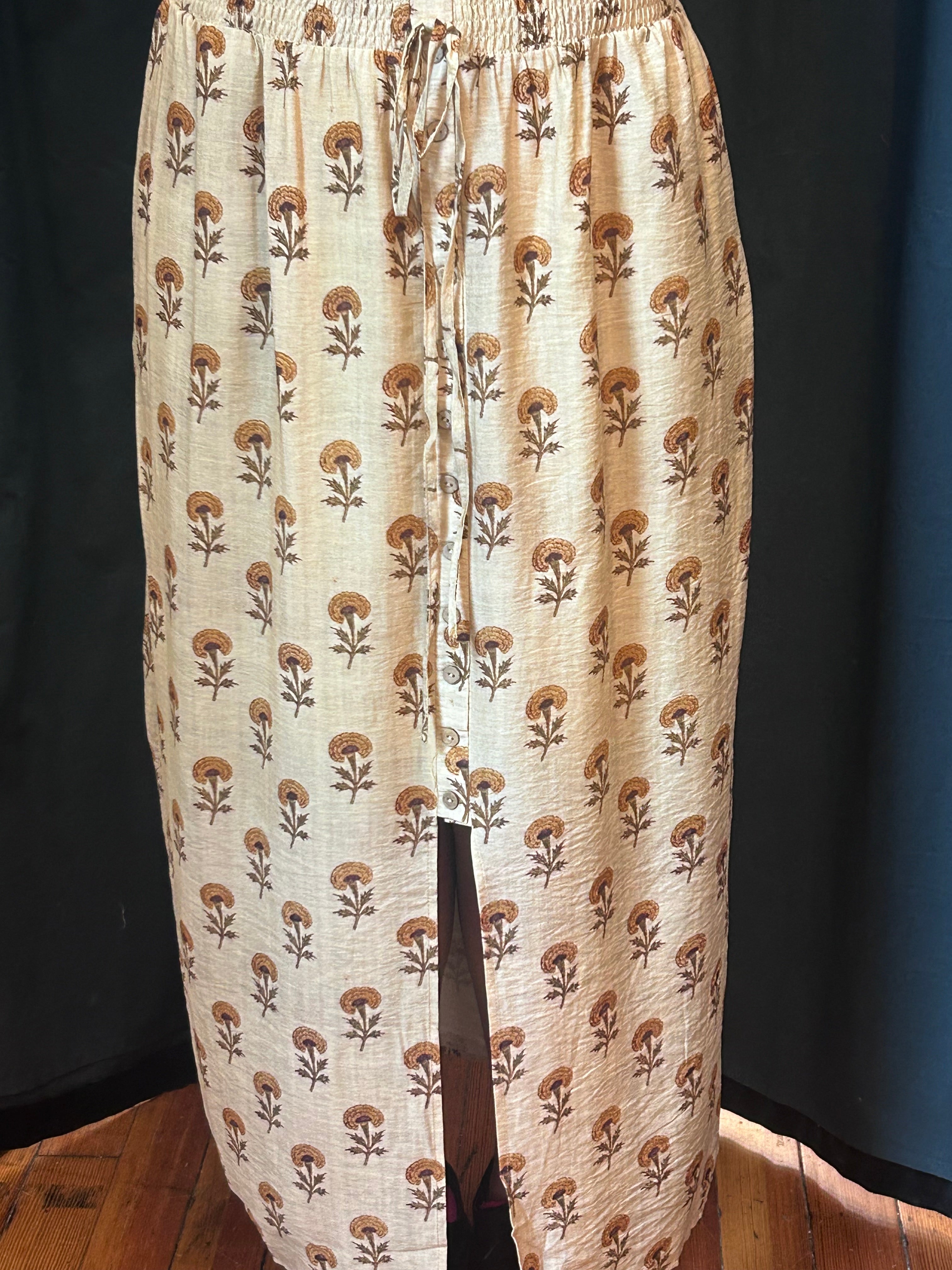 A beige maxi skirt with a brown and tan floral block print pattern, featuring an elastic waistband and a front slit, displayed on a mannequin against a dark background.